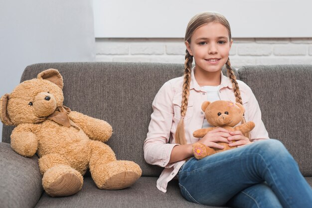 Ritratto di una ragazza sorridente che si siede con il piccolo orsacchiotto sul sofà grigio