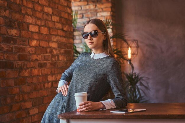 Ritratto di una ragazza sorridente con occhiali da sole vestita con un elegante abito grigio tiene una tazza di caffè da asporto e tablet mentre si appoggia sul tavolo in una stanza con interni soppalcati.