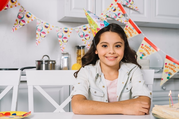 Ritratto di una ragazza sorridente di compleanno che porta il cappello del partito sulla testa che guarda l&#39;obbiettivo