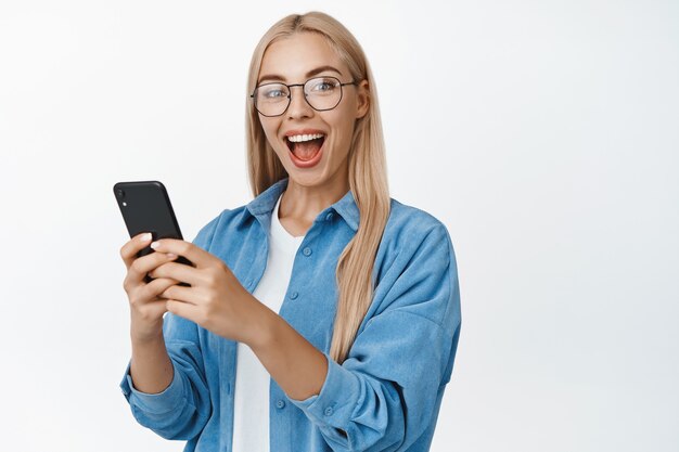 Ritratto di una ragazza sorridente eccitata che tiene in mano il telefono cellulare e reagisce stupita, con gli occhiali, in piedi sul bianco.