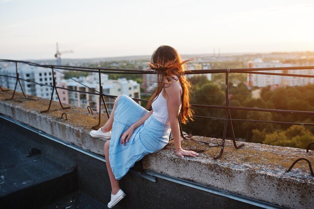 Ritratto di una splendida giovane donna in maglietta bianca e gonna blu seduta sul bordo dell'edificio al tramonto