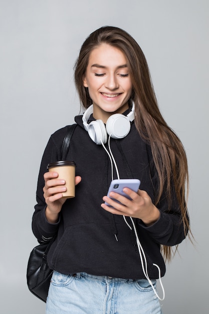 Ritratto di una studentessa attraente allegra con lo zaino che ascolta la musica con le cuffie mentre mostrando il telefono cellulare dello schermo in bianco e ballare isolato sopra la parete bianca