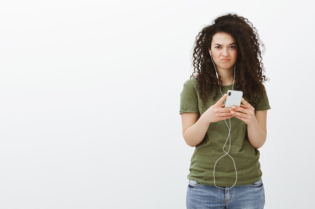 Ritratto di una studentessa attraente scontenta per nulla impressionato con capelli castani ricci, tenendo lo smartphone e indossando gli auricolari, guardando dispiaciuto e triste