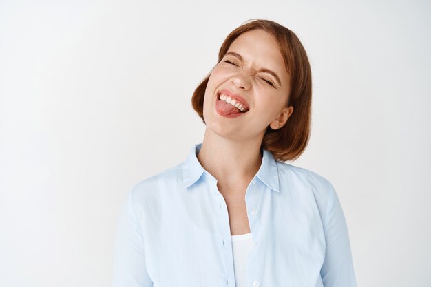 Ritratto di una studentessa spensierata che sorride con gli occhi chiusi, che mostra la lingua e che ha la lingua, in piedi giocoso sul muro bianco