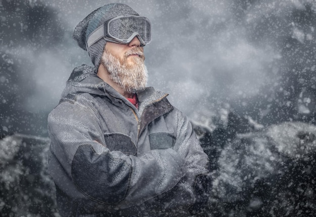 Ritratto di uno snowboarder rosso con la barba piena in un cappello invernale e occhiali protettivi vestito con un cappotto da snowboard in posa sullo sfondo delle montagne.