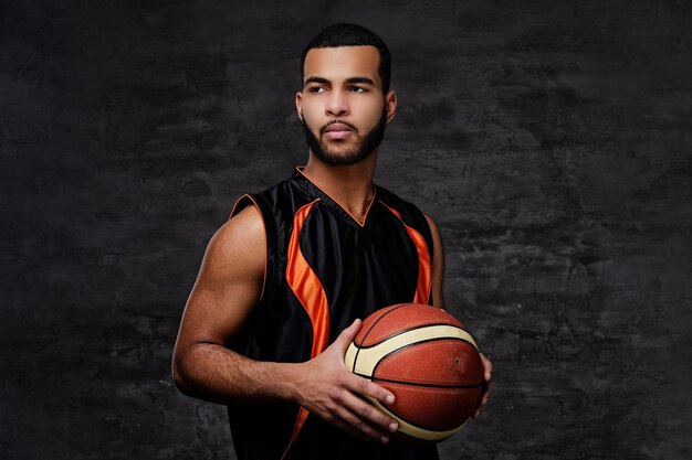 Ritratto di uno sportivo afroamericano. Giocatore di basket in abbigliamento sportivo con una palla su sfondo scuro.