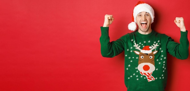 Ritratto di uomo allegro e attraente che celebra il nuovo anno indossando un maglione verde e un cappello da Babbo Natale che grida di gioia vincendo o trionfando in piedi su sfondo rosso