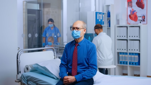 Ritratto di uomo anziano che indossa la maschera all'appuntamento dal medico, seduto sul letto d'ospedale in attesa dei risultati di COVID-19. Sistema di medicina meccanica sanitaria durante la pandemia globale, ripresa al rallentatore portatile