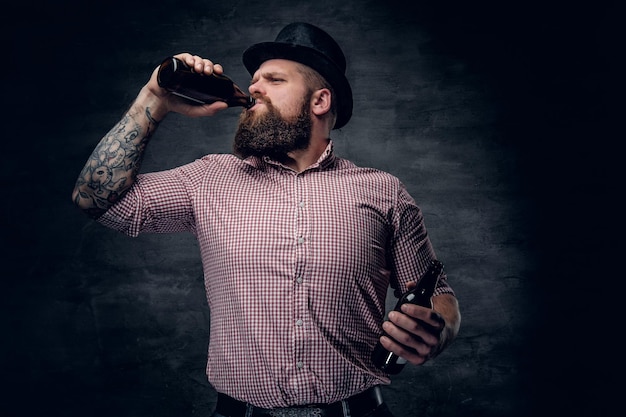 Ritratto di uomo barbuto con una camicia a quadri e un cappello a cilindro, che beve birra da una bottiglia.