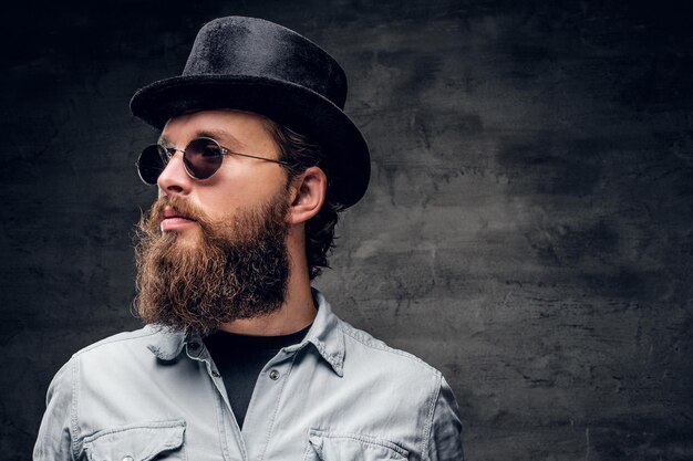 Ritratto di uomo barbuto curato in cappello e occhiali da sole su sfondo scuro.