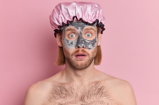 Ritratto di uomo barbuto sorpreso guarda scioccato indossa cappello da doccia applica maschera di argilla nutriente per il ringiovanimento e la riduzione delle rughe si leva in piedi con le spalle nude.