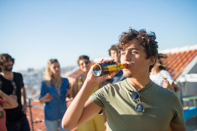 Ritratto di uomo bello con i capelli ricci alla festa. Giovane ragazzo caucasico in maglietta verde che beve birra. Amici di diverse nazionalità in background. Festa, concetto di amicizia