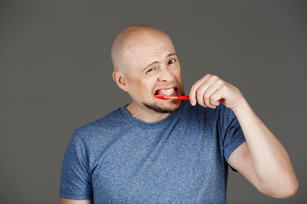 Ritratto di uomo bello divertente in camicia grigia lavarsi i denti sopra il muro scuro