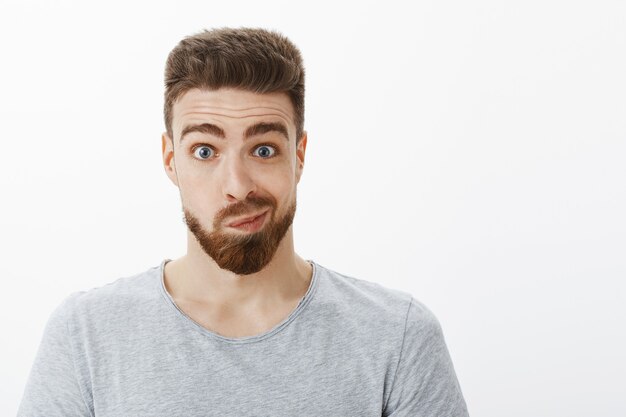 Ritratto di uomo bello sciocco e divertente con barba, baffi e occhi azzurri sorridendo facendo un viso goffo incerto guardandosi allo specchio e pensando di apportare modifiche in posa contro il muro grigio