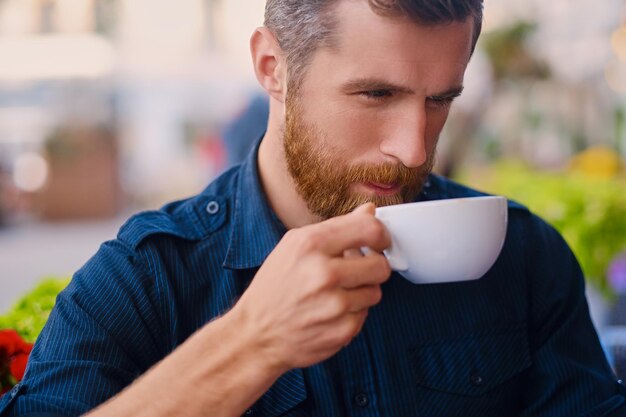 Ritratto di uomo casual rosso barbuto beve caffè in un caffè in una strada.