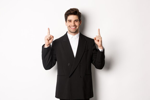 Ritratto di uomo caucasico attraente in elegante abito nero, puntando le dita in alto e sorridente, mostrando la pubblicità di natale, in piedi su sfondo bianco.