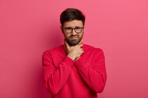 Ritratto di uomo caucasico barbuto infelice si strozza, non riesce a respirare, soffoca e tiene le mani sul collo, aggrotta le sopracciglia, soffre di mal di gola e attacchi asmatici, posa al coperto, vestito casualmente