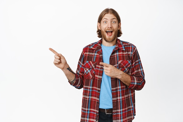 Ritratto di uomo caucasico eccitato con barba e faccia sorridente felice, che punta il dito a sinistra, mostrando sconti incredibili, grande promozione, in piedi su sfondo bianco