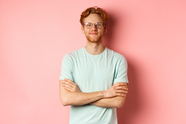 Ritratto di uomo caucasico soddisfatto con capelli e barba rossi, braccia incrociate sul petto e sorridente con faccia compiaciuta, con gli occhiali, in piedi su sfondo rosa.