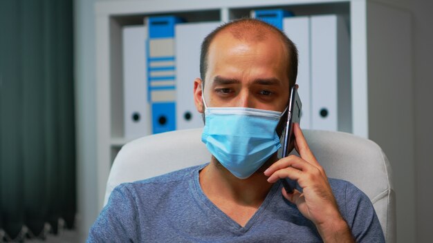 Ritratto di uomo con maschera protettiva che parla al telefono con i partner nella stanza moderna dell'ufficio durante il covid-19. Libero professionista che lavora nel nuovo normale posto di lavoro chattando scrivendo parlando su smartphone.