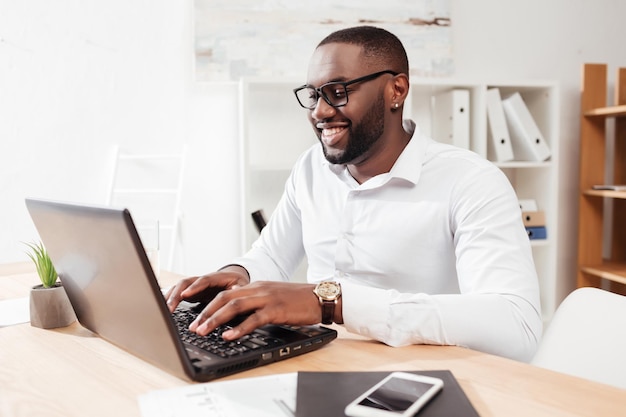 Ritratto di uomo d'affari afroamericano sorridente in camicia bianca e occhiali seduto e lavorando sul suo computer portatile in ufficio isolato