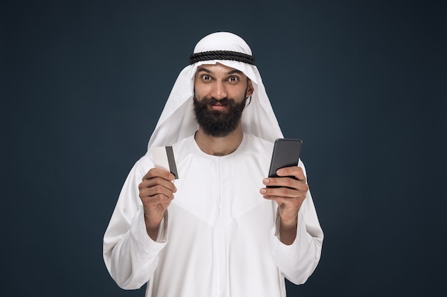 Ritratto di uomo d'affari arabo saudita su sfondo blu scuro per studio. Uomo che utilizza smartphone per pagare la bolletta, acquisti online o scommesse. Concetto di affari, finanza, espressione facciale, emozioni umane.