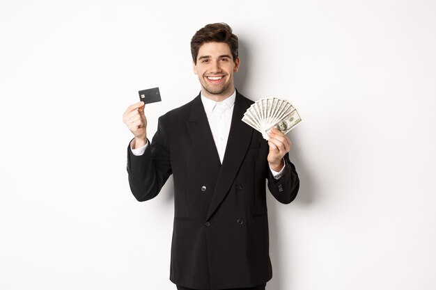 Ritratto di uomo d'affari attraente in abito nero, mostrando soldi e carta di credito, sorridente soddisfatto, in piedi su sfondo bianco.