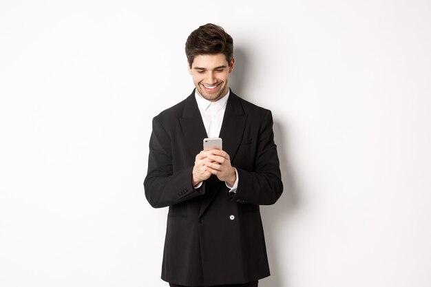 Ritratto di uomo d'affari bello ed elegante in abito nero, scrivendo un messaggio, sorridendo e guardando smartphone, in piedi su sfondo bianco.