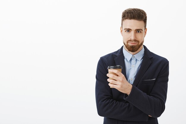 Ritratto di uomo d'affari bello fiducioso e di successo in abito elegante che tiene tazza di caffè di carta sorridendo con gioia comando di simpatia e tenendo sotto controllo gli affari contro il muro bianco