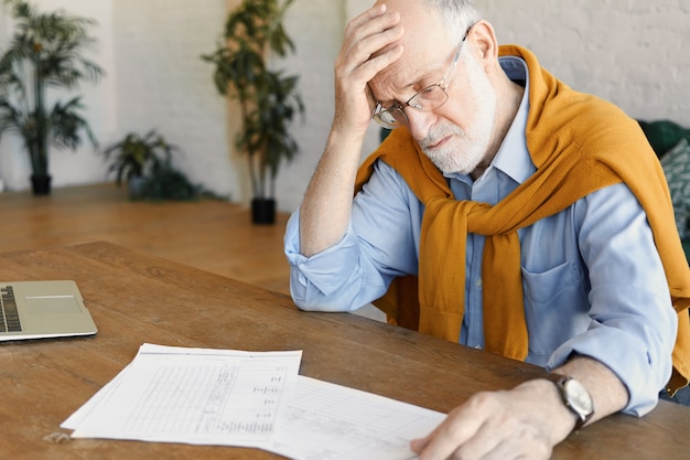 Ritratto di uomo d'affari caucasico maturo stressato in abiti formali e occhiali seduto davanti al computer portatile aperto, studiando documenti, affrontando problemi finanziari, tenendo la mano sulla sua testa calva