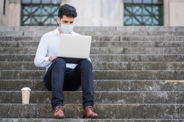 Ritratto di uomo d'affari che indossa la maschera per il viso e utilizzando il suo laptop mentre è seduto sulle scale all'aperto