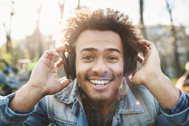 Ritratto di uomo dalla carnagione scura adulto positivo che sorride ampiamente mentre è seduto nel parco, ascoltando la musica in cuffia e tenendoli con le mani per sentire meglio.