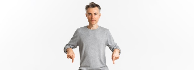 Ritratto di uomo di mezza età di successo in maglione grigio che punta le dita verso il basso e guardando fiducioso alla macchina fotografica che mostra il logo in piedi su sfondo bianco