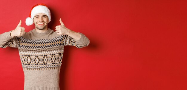 Ritratto di uomo felice attraente con cappello da Babbo Natale e maglione che mostra pollice in su in approvazione e sorridente augurando buon Natale in piedi su sfondo rosso