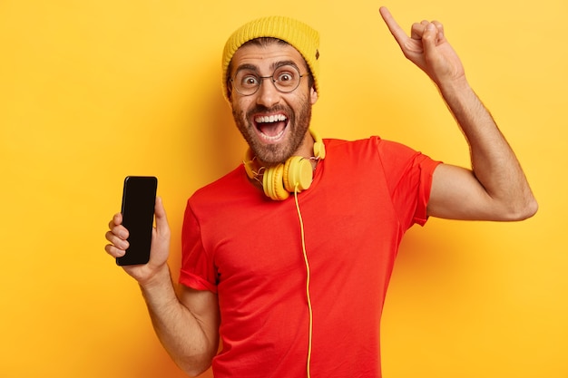 Ritratto di uomo felice con la barba lunga tiene il telefono cellulare con lo schermo vuoto, alza il braccio e indica con il dito indice sopra, ha un'espressione facciale felice, indossa un cappello giallo e una maglietta rossa, usa le cuffie