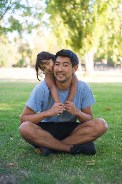 Ritratto di uomo felice e bambina sul campo erboso nel parco. Padre asiatico seduto sull'erba e la sua bella figlia in piedi dietro di lui entrambi guardando la fotocamera. Concetto di vacanze estive e genitorialità