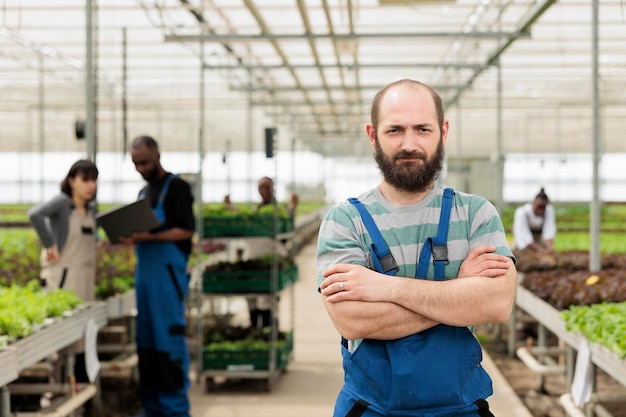 Ritratto di uomo fiducioso che lavora in serra mentre gli ingegneri agricoli utilizzano il laptop per determinare i tassi di crescita. Uomo caucasico in piedi nella piantagione di microgreens idroponica.