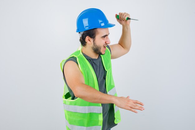 Ritratto di uomo giovane costruttore alzando la mano per colpire con un cacciavite in uniforme e guardando folle vista frontale