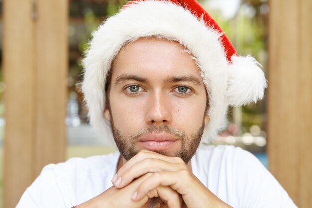 Ritratto di uomo giovane e bello hipster con stoppie e sorriso fiducioso in posa all'interno, tenendo le mani giunte, indossando il cappello di Babbo Natale rosso con pelliccia bianca