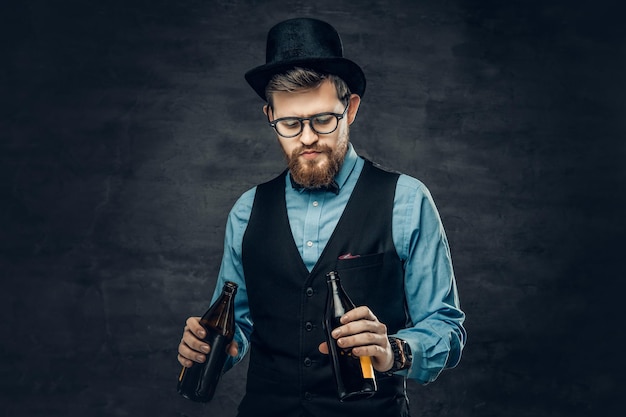 Ritratto di uomo hipster barbuto vestito con una camicia blu, un gilet elegante e un cappello a cilindro tiene due bottiglie di birra artigianale con un pensiero a una festa.