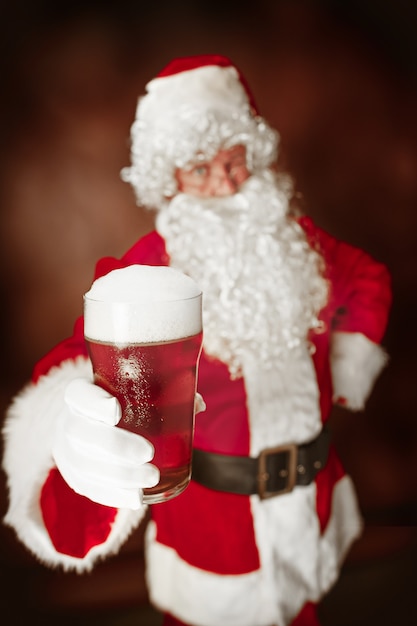 Ritratto di uomo in costume da Babbo Natale - con una lussuosa barba bianca, cappello di Babbo Natale e un costume rosso su sfondo rosso in studio con birra