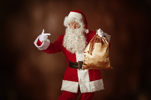 Ritratto di uomo in costume da Babbo Natale - con una lussuosa barba bianca, cappello di Babbo Natale e un costume rosso su sfondo rosso in studio con regali