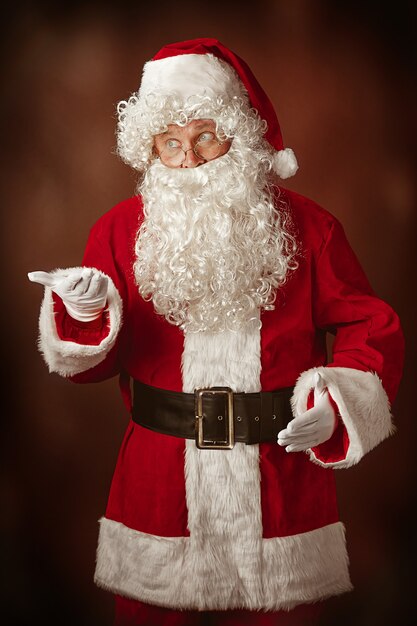 Ritratto di uomo in costume da Babbo Natale - con una lussuosa barba bianca, cappello di Babbo Natale e un costume rosso su sfondo rosso per studio