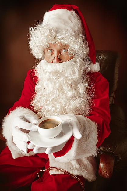 Ritratto di uomo in costume da Babbo Natale - con una lussuosa barba bianca, cappello di Babbo Natale e un costume rosso su sfondo rosso studio seduto su una sedia con una tazza di caffè