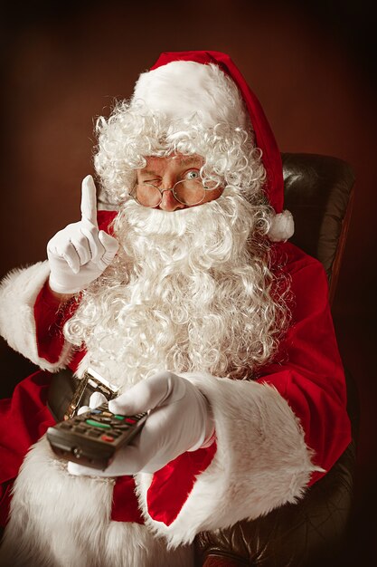 Ritratto di uomo in costume di Babbo Natale con una lussuosa barba bianca, cappello di Babbo Natale e un costume rosso al rosso seduto su una sedia con telecomando TV