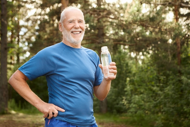 Ritratto di uomo in pensione caucasico attivo gioioso con barba e testa in grassetto tenendo la mano sulla sua vita e bere acqua fresca dalla bottiglia di vetro, avendo riposo dopo l'allenamento fisico mattutino nel parco