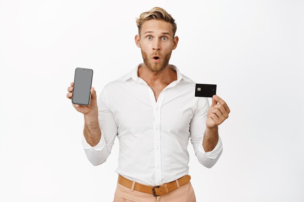 Ritratto di uomo in possesso di carta di credito e che mostra lo schermo del telefono cellulare rendendo l'espressione del viso sorpreso WOW sguardo in piedi su sfondo bianco