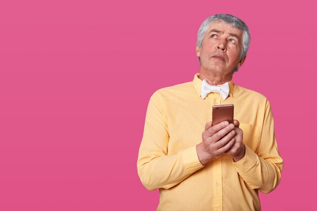 Ritratto di uomo maturo con rughe e capelli grigi, vestito in camicia gialla e farfallino bianco, tenendo smartphone nelle mani, cerca. Uomo anziano con il telefono cellulare che posa negli isolati dello studio sul rosa.