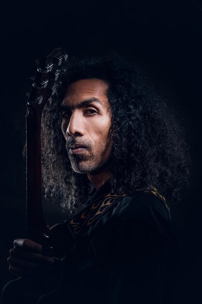 Ritratto di uomo riccio attraente con la chitarra in studio fotografico scuro.