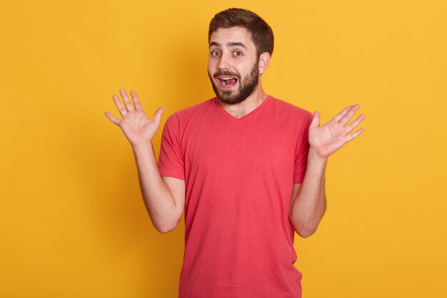 Ritratto di uomo sorpreso, bel maschio allargando le mani, in posa isolato sul muro giallo, attraente con la barba lunga ragazzo che indossa maglietta casual rossa. Il concetto di emozioni umane.